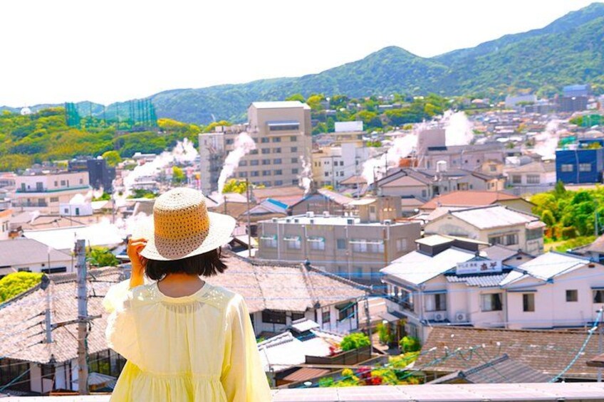 A person in a yellow dress and straw hat overlooking a cityscape with buildings and mountains in the background. 2