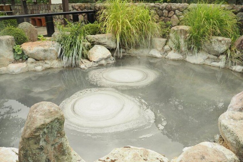 A hot spring with circular ripples in the water, surrounded by rocks and greenery. 5