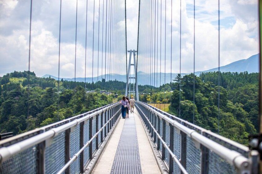 A suspension bridge with a metal railing, surrounded by lush green forest and distant mountains. 1