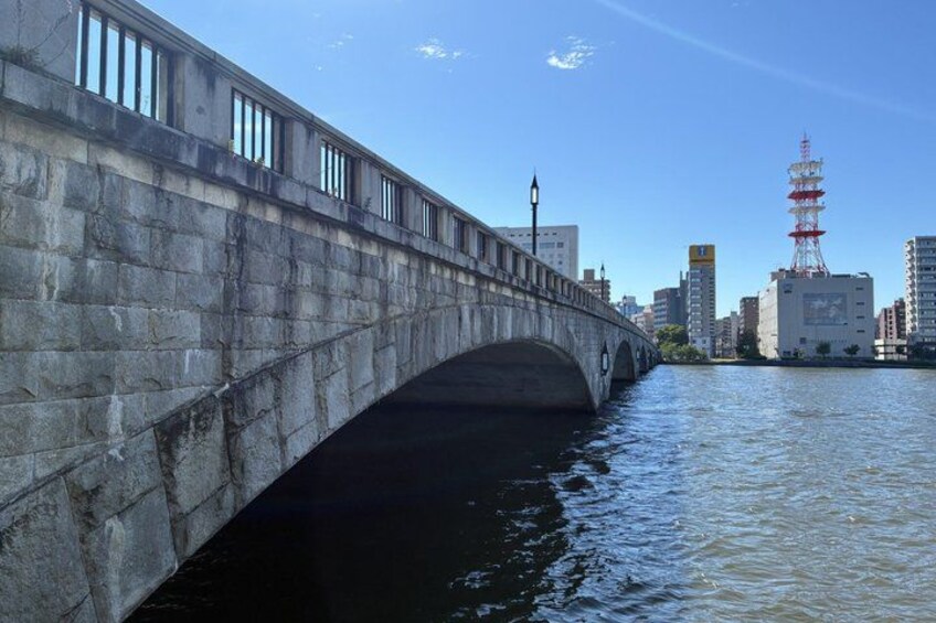 A stone bridge over water with buildings and a red tower in the background. 3