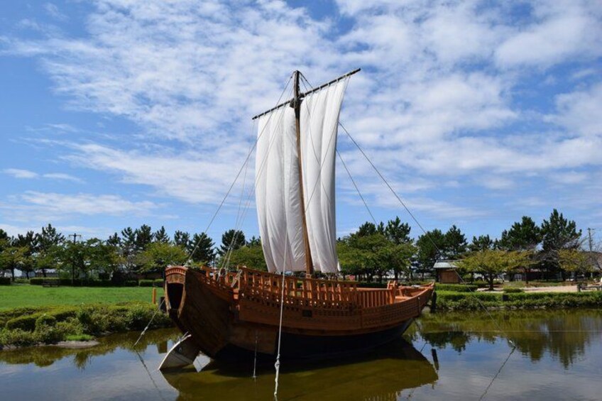 A wooden boat with white sails docked in a calm body of water, surrounded by greenery and trees. 4