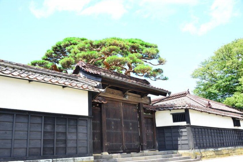 A traditional Japanese building with a tiled roof, a large wooden gate, and a prominent pine tree. 2