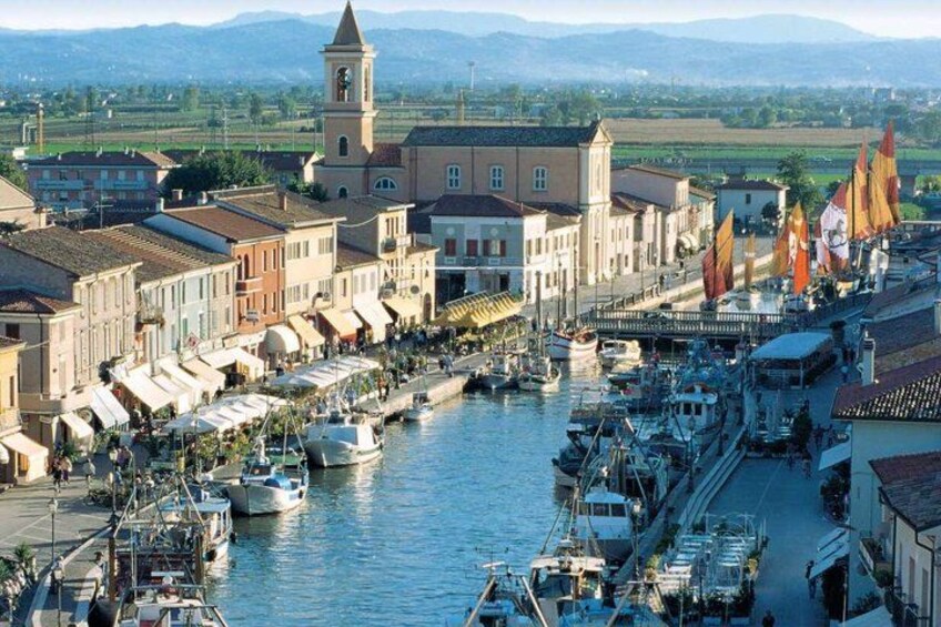 A riverside town with boats docked along the water, buildings, and a tower. 3