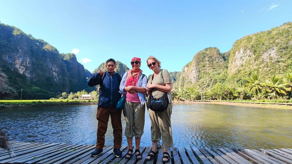 Three people standing on a wooden dock by a river with a mountainous landscape in the background. 4