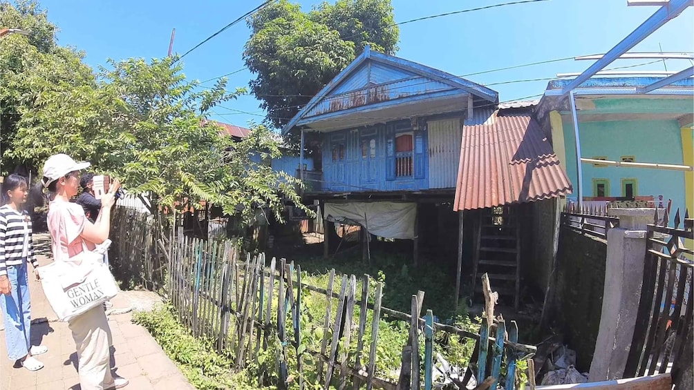 A blue house with a corrugated metal roof, surrounded by a wooden fence and greenery. 1