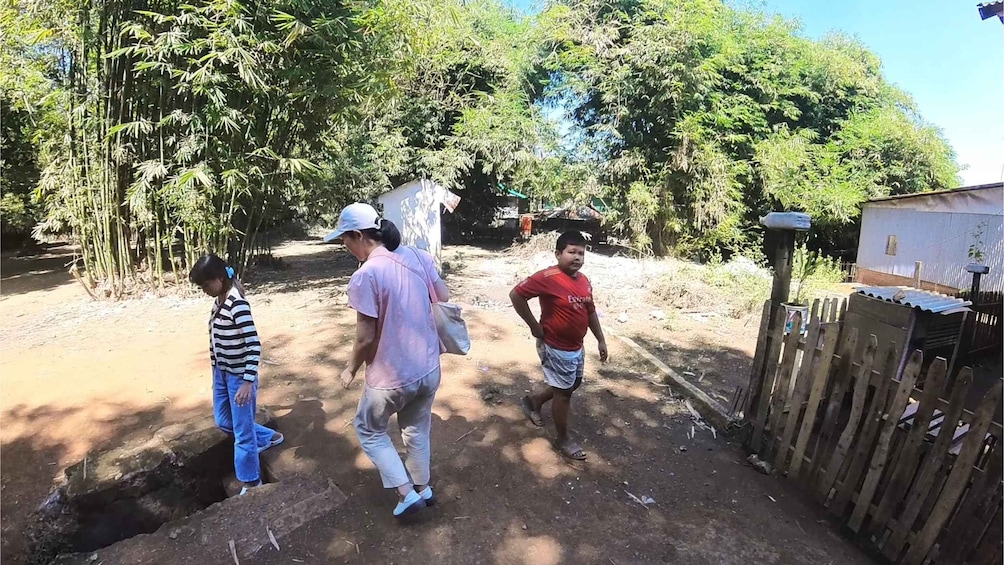Three people standing in a rural setting with a dirt path, bamboo trees, and a wooden fence. 2