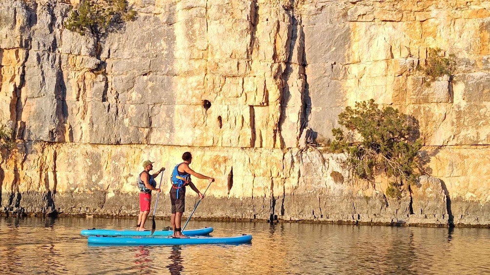 Two people paddleboarding on a calm river with a rocky cliff in the background. 4