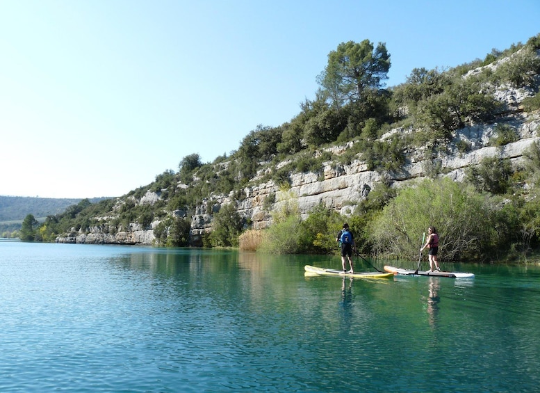 Two people paddleboarding on a calm lake with a rocky hillside in the background. 1