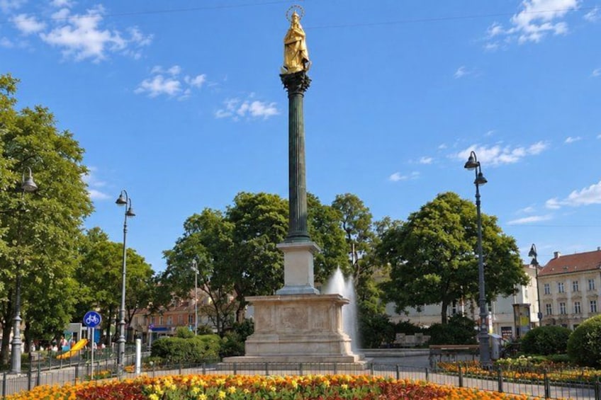 A tall monument with a statue on top, surrounded by trees and a flower bed. 2