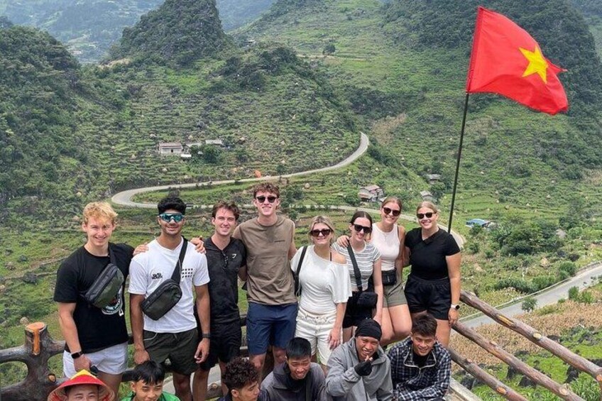 A group of people standing on a hillside with a flag, surrounded by lush green hills and a winding road. 1