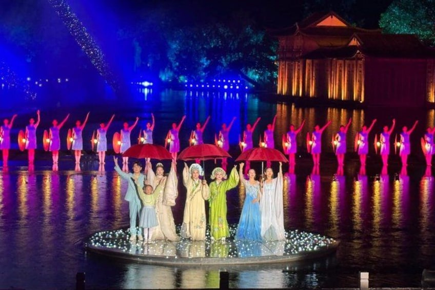 A nighttime outdoor performance with dancers in colorful costumes on a stage by a body of water, illuminated by blue and purple lights. 3