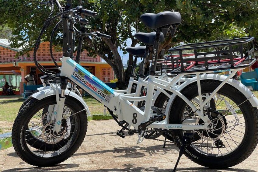 A white electric bicycle with a front basket and a rear rack, parked on a sandy surface with trees and a building in the background. 1