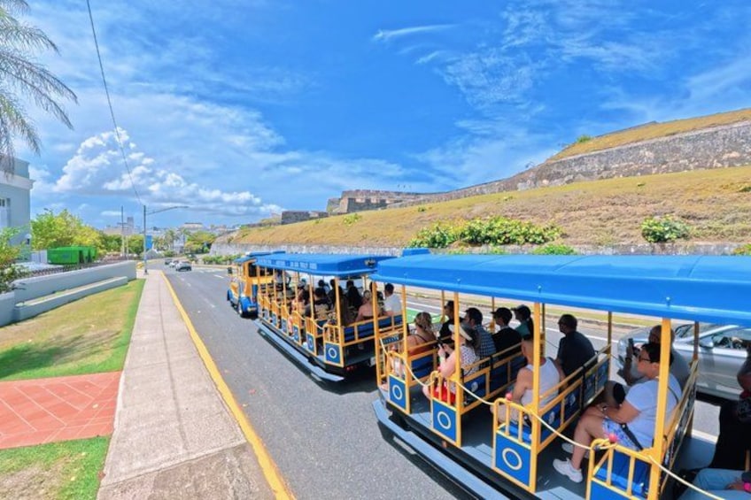 A tram with passengers, a road, a grassy area, and a hilly landscape. 1
