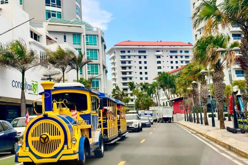 A yellow and blue train on a city street with palm trees and buildings in the background. 4