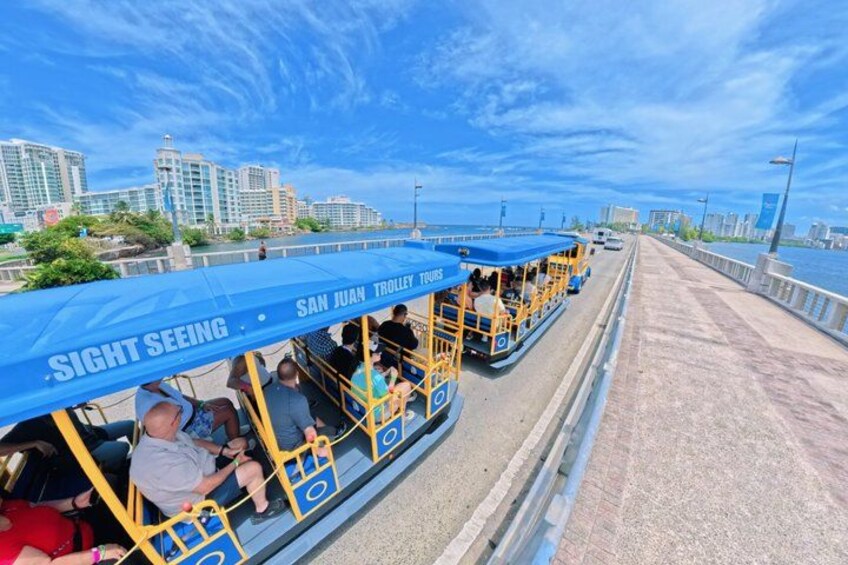 A San Juan trolley with passengers, a bridge, and city buildings in the background. 3