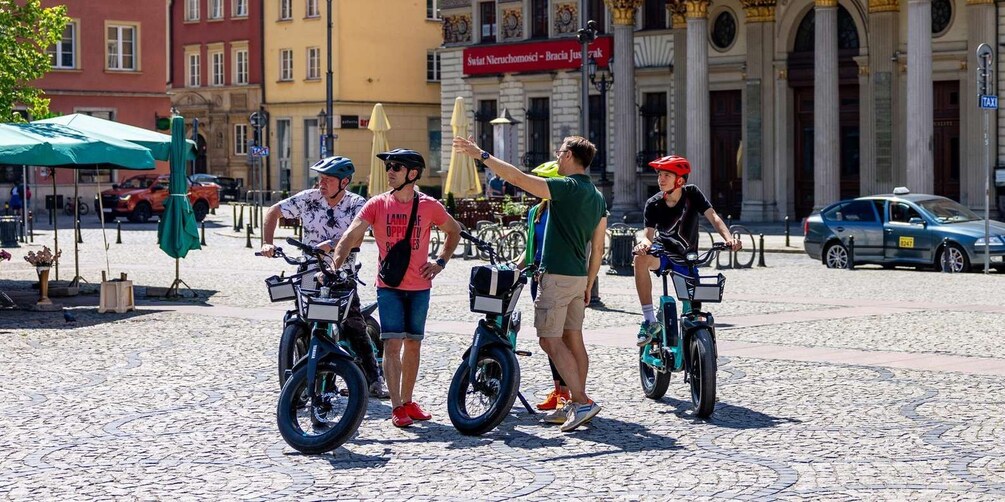 Four people on electric unicycles on a cobblestone street with buildings and a taxi in the background. 3