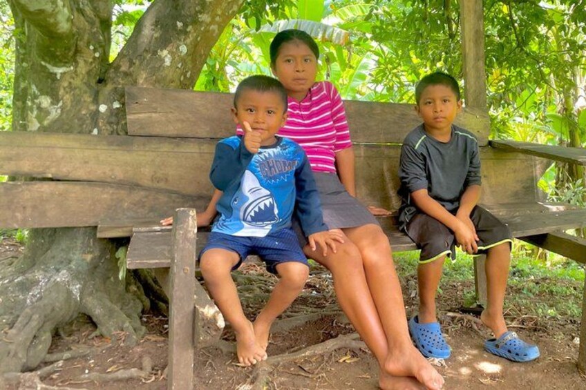 Three children sitting on a wooden bench in a natural setting with trees and foliage in the background. 1