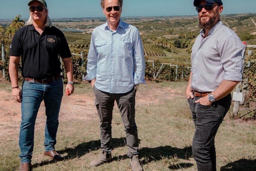 Three men standing on a grassy hill with vineyards in the background. 3