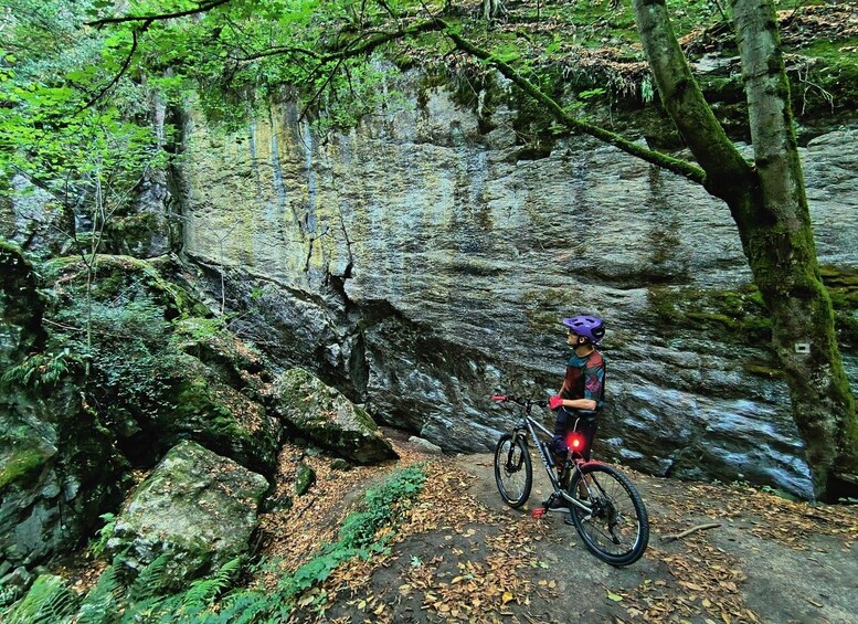 A mountain biker on a trail with a rocky cliff and dense forest. 1