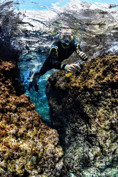 A diver in a black wetsuit with yellow accents, underwater near rocky formations with clear water. 2