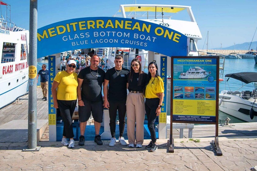 A group of people standing under a sign for Mediterranean One Glass Bottom Boat Blue Lagoon Cruises. 4
