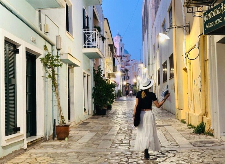 A narrow street with buildings on either side, a person walking away from the camera, and a church with a dome in the background. 1