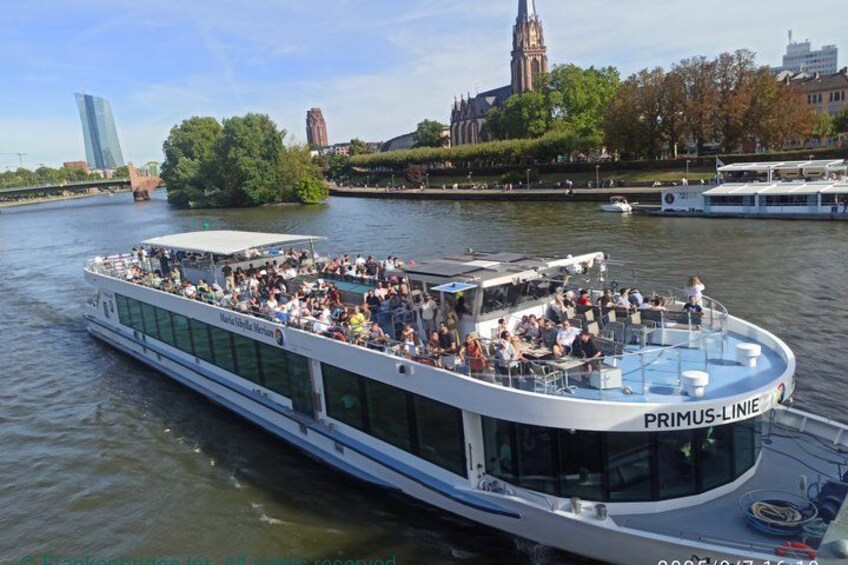 A river cruise boat named PRIMUS-LINIE with passengers, a cityscape with a church and modern buildings in the background. 4