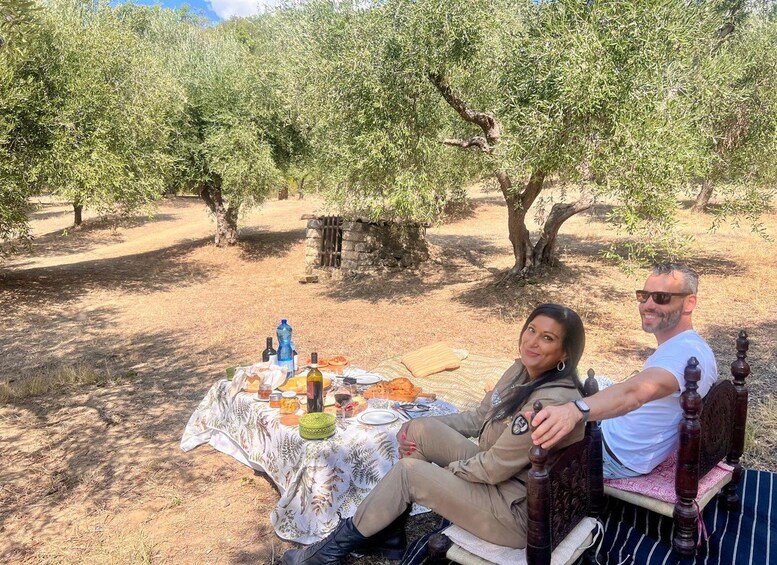 Two people sitting on a bench in an olive grove with a picnic setup on the ground. 2