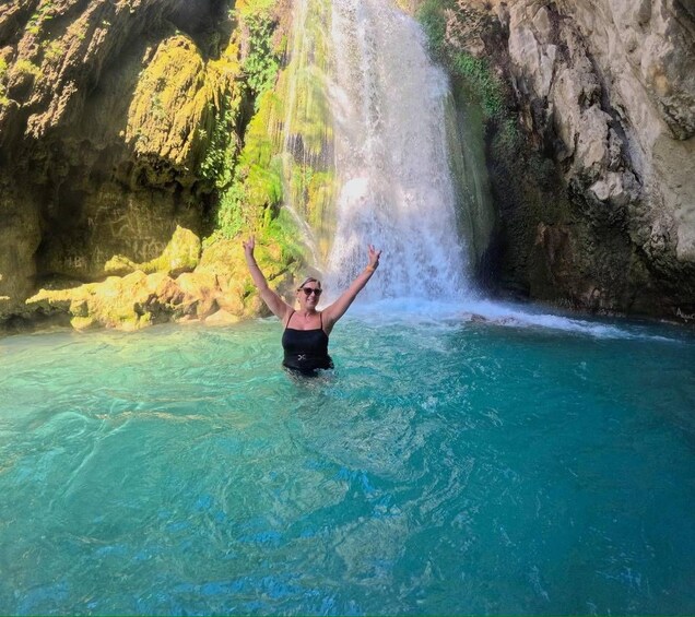 A person in a black swimsuit standing in clear blue water with a waterfall in the background. 1