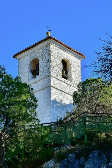 A white bell tower with two arched openings, surrounded by greenery and a clear blue sky. 3