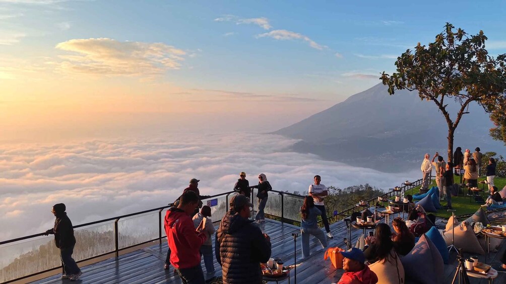 A group of people on a wooden platform overlooking a mountainous landscape with a view of clouds below and a volcano in the distance. 1