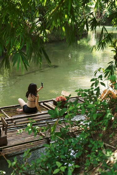 A person sitting on a bamboo raft in a calm river surrounded by lush greenery. 1