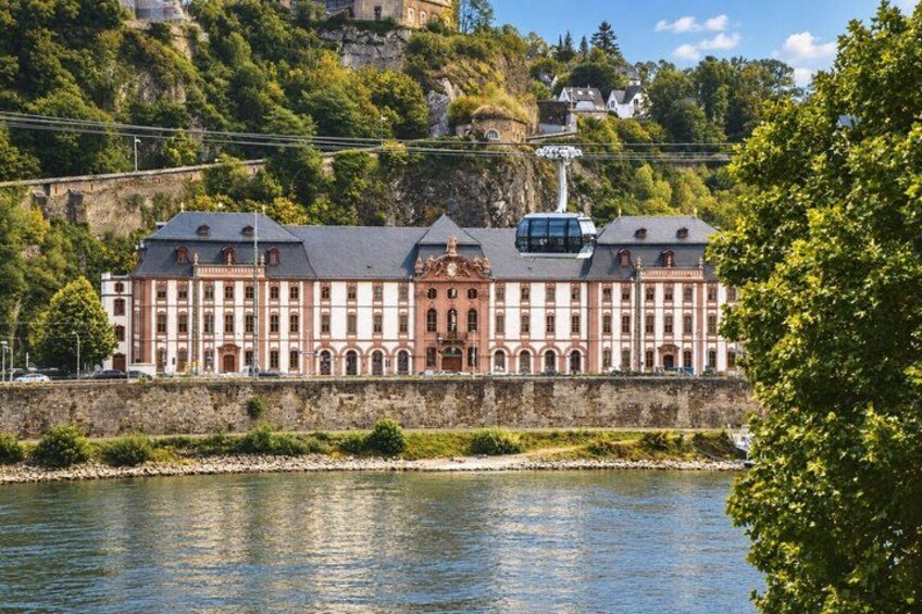 A large, historic building with a red facade, situated by a river with a rocky hillside in the background. 3