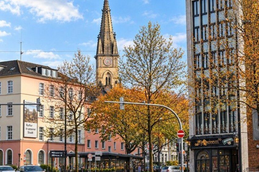 A city street with autumn trees, a church tower, and modern buildings. 2
