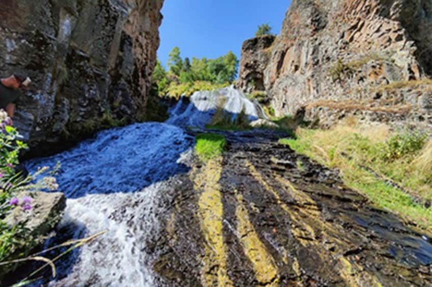 Una cascada se precipita por un acantilado rocoso, rodeada de vegetación y un cielo azul despejado. 1