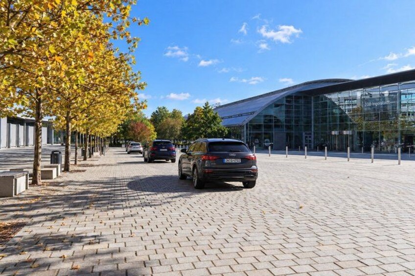 A paved walkway with parked cars, a modern building with glass facade, and trees with yellow leaves. 2