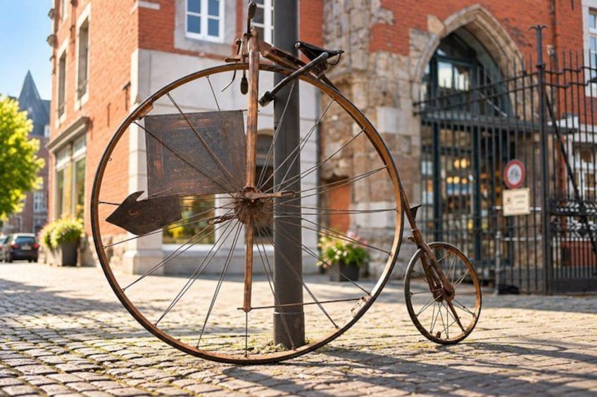 Un vélo ancien, doté d’une grande roue avant et d’une plus petite roue arrière, appuyé contre un poteau sur une rue pavée. 3
