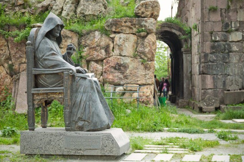 A statue of a robed figure seated on a stone bench, with a stone archway and greenery in the background. 2