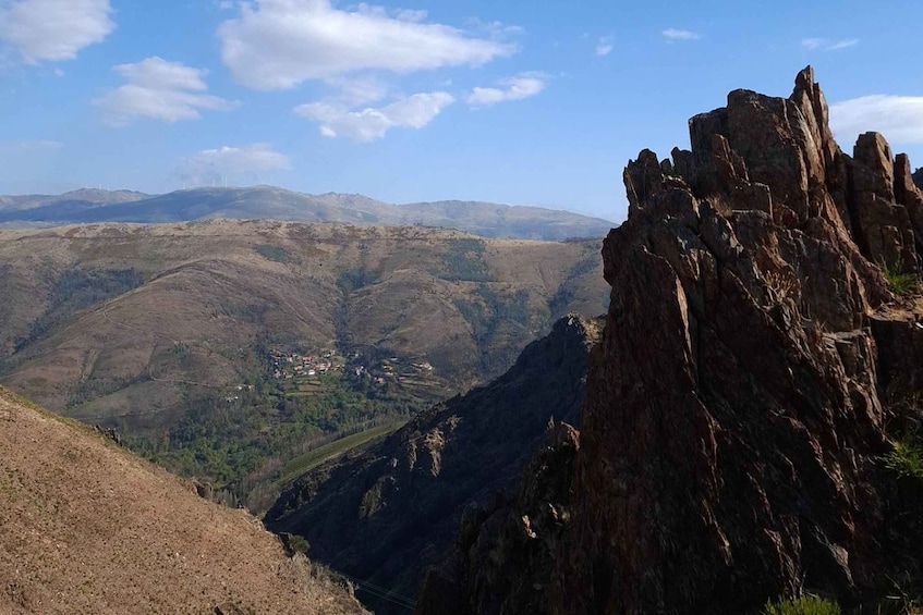 A mountainous landscape with a rocky outcrop in the foreground and a valley with scattered buildings in the background. 1