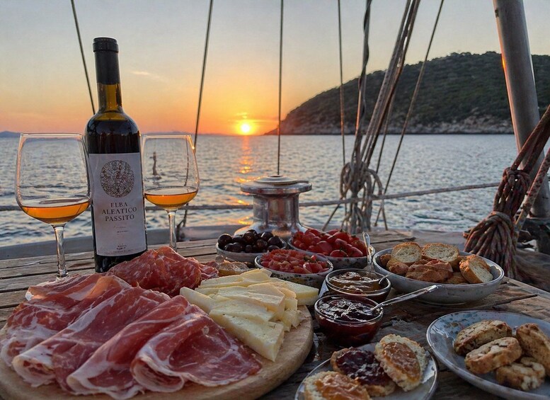 A sunset view from a boat with a bottle of Elba Aleatico Passito wine, two glasses, and a variety of cheeses, meats, olives, and bread. 2