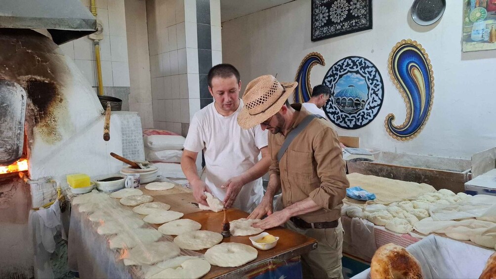 Two men preparing dough on a table in a kitchen with a traditional oven and decorative wall plates. 1