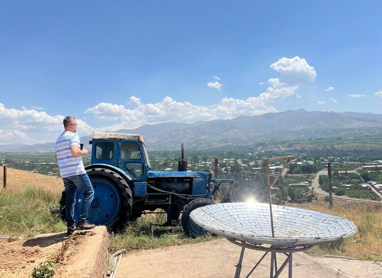 A man standing next to a blue tractor on a dirt path with a solar panel in the foreground and a valley with scattered houses in the background. 3