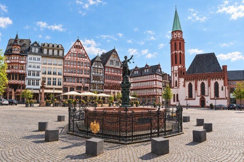A cobblestone square with a fountain, historic buildings, and a church tower. 2