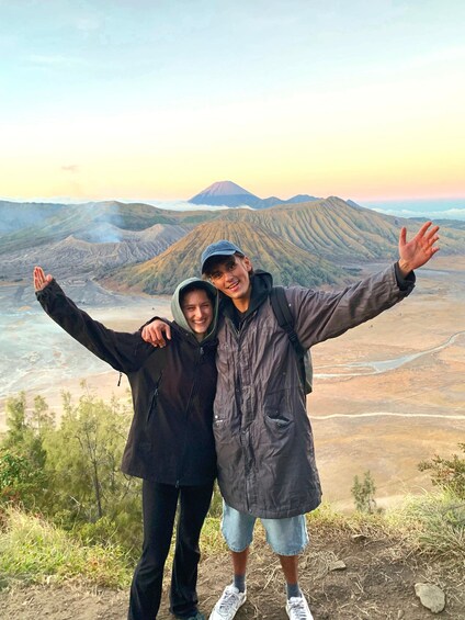 Two people standing with arms outstretched, overlooking a mountainous landscape with a river and a volcano in the background. 1