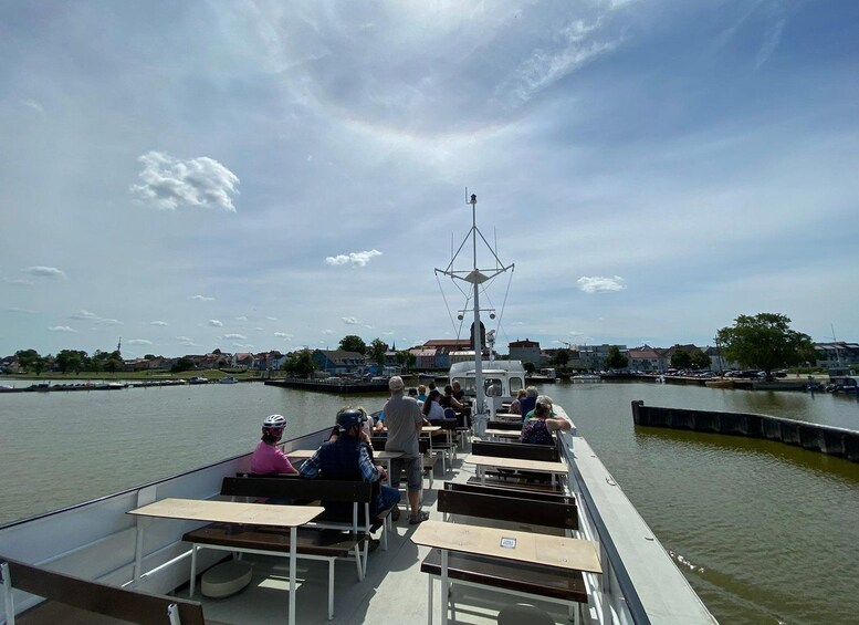 Un bateau avec des personnes assises à des tables, amarré dans une marina, avec des bâtiments et un ciel dégagé en arrière-plan. 2