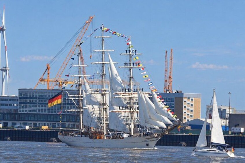 A large sailing ship with multiple masts and flags, docked at a port with cranes and buildings in the background. 5