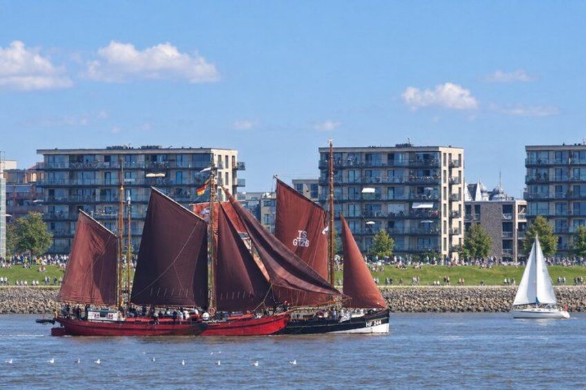 A red sailboat with multiple sails on a body of water, a white sailboat in the background, and residential buildings along the shore. 4