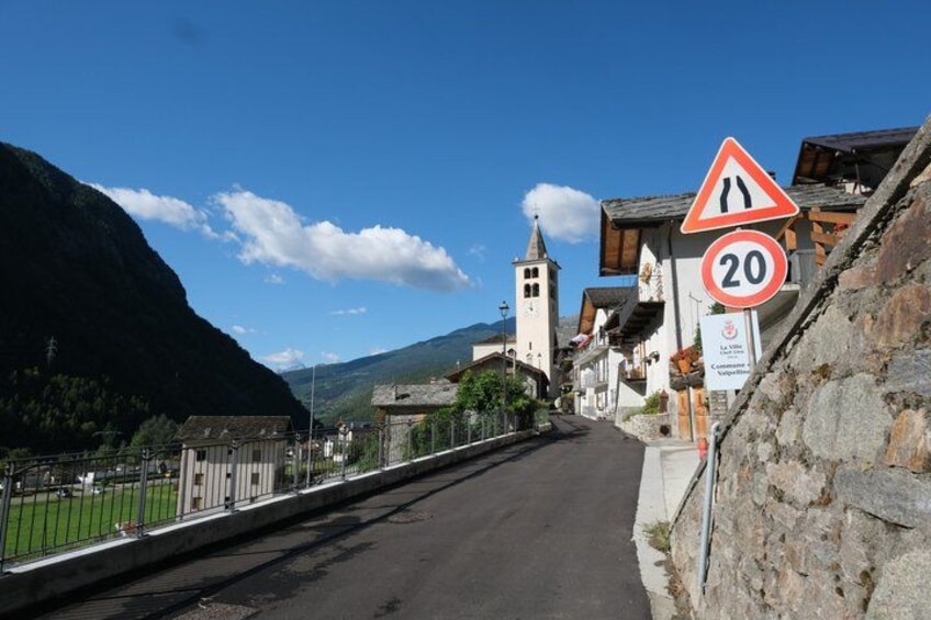 A narrow mountain road with a 20 km/h speed limit sign, a narrow road warning sign, and a church with a bell tower. 3