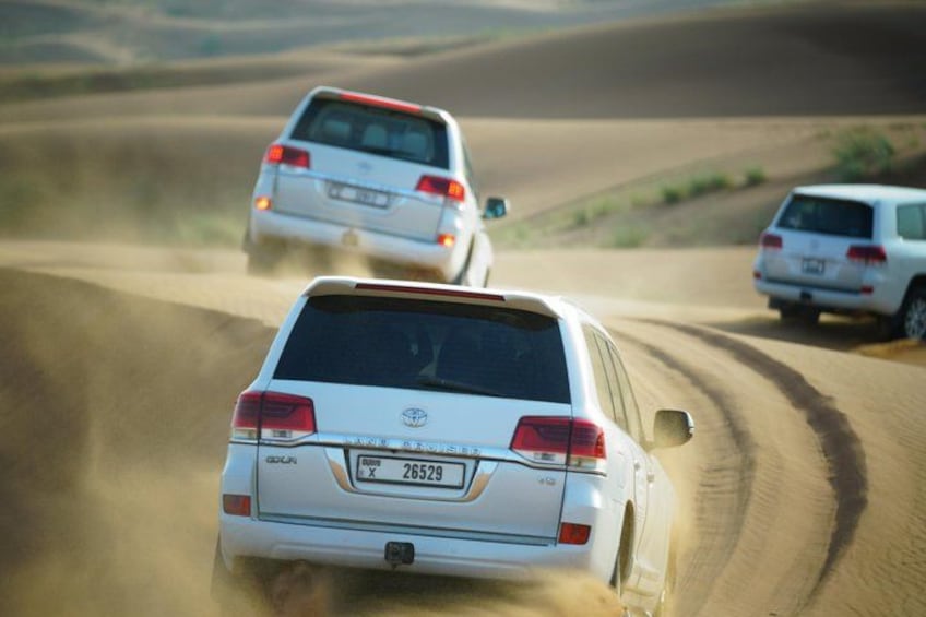 Two white SUVs driving on a dusty road in a desert landscape. 1