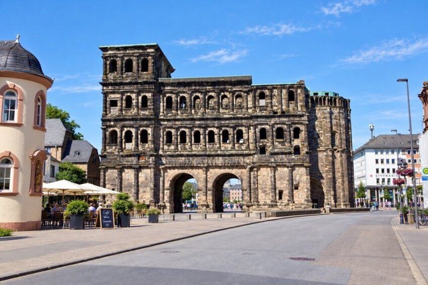 A historic stone archway with multiple arches and a tower, surrounded by modern buildings and a clear sky. 2
